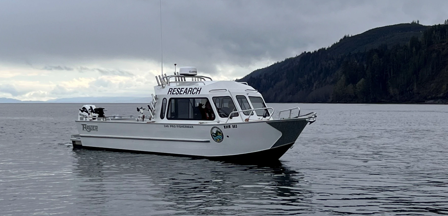 Research vessel labeled '240 Pro Fisherman' on calm waters, featuring the WDFW logo on its side. The boat is equipped with antennas and fishing equipment, set against a backdrop of cloudy skies and forested hills.